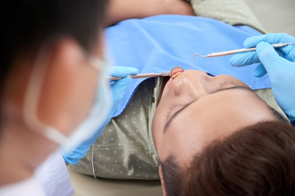 A patient lying back in a dental chair with a dentist examining their teeth using dental instruments.
